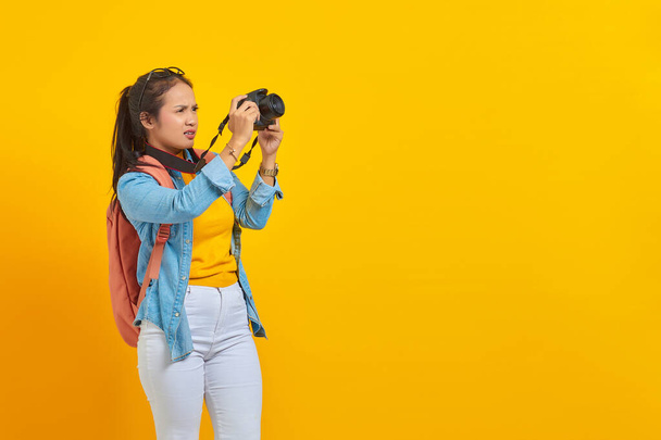 Portrét překvapené mladé asijské ženy v džínových šatech s batohem fotografování na fotoaparátu profesionální izolované na žlutém pozadí - Fotografie, Obrázek