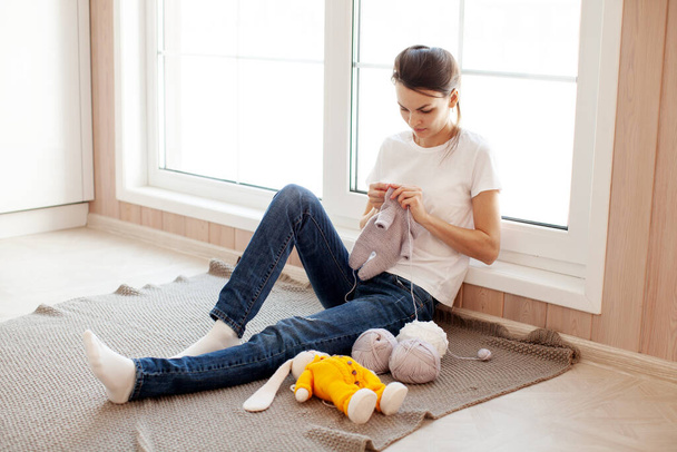 Frau strickt Kleidung für Spielzeug-Osterhasen, die im weißen Hausinneren neben einem hellen riesigen Fenster sitzt. Hobby und Basteln oder Handwerk - Foto, Bild