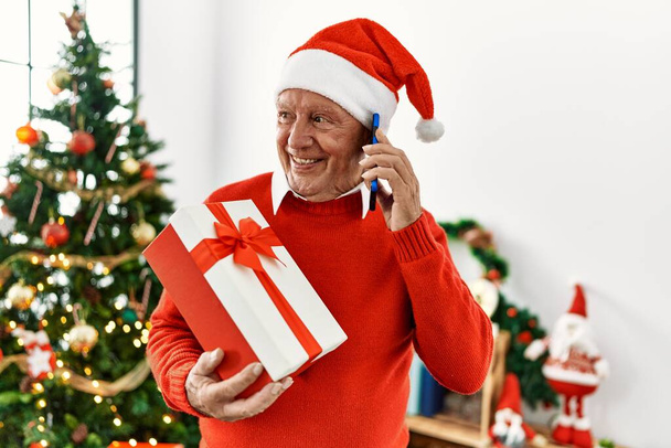 Старший седой мужчина разговаривает на смартфоне, держа рождественский подарок дома. - Фото, изображение