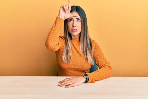 Jonge Latijns-Amerikaanse vrouw in casual kleding zittend op de tafel lachen van mensen met vingers op het voorhoofd doen verliezer gebaar bespotten en beledigen.  - Foto, afbeelding