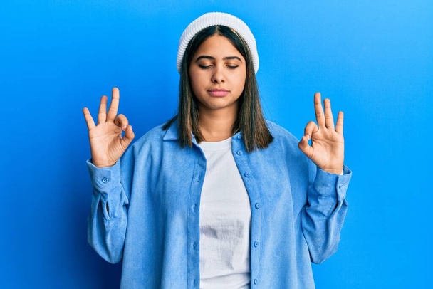 Jonge latijnse vrouw draagt leuke wollen pet ontspannen en glimlachend met gesloten ogen meditatie gebaar met vingers. yoga concept.  - Foto, afbeelding