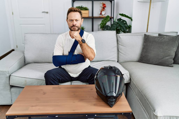 Middle age man with beard wearing arm on sling because motorbike accident serious face thinking about question with hand on chin, thoughtful about confusing idea  - Photo, Image