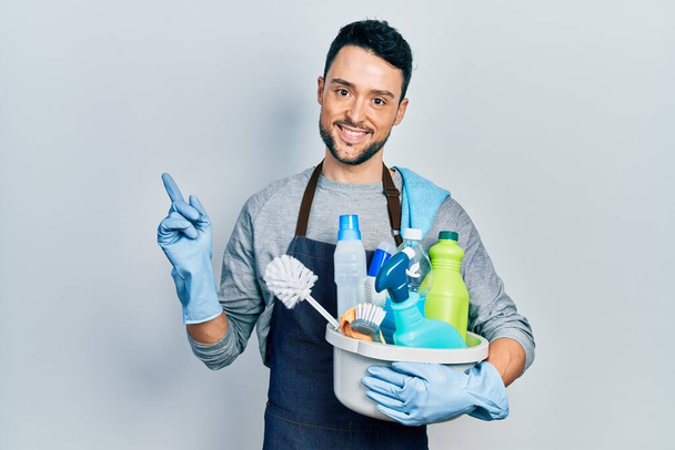 Молодой латиноамериканец держит чистящие средства, улыбаясь счастливо указывая рукой и пальцем в сторону  - Фото, изображение