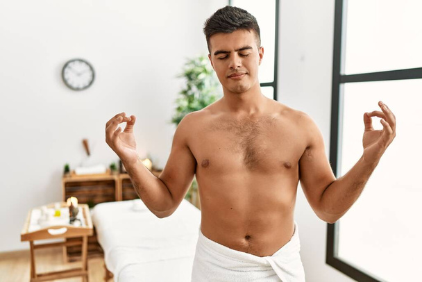 Young hispanic man standing shirtless at spa center relax and smiling with eyes closed doing meditation gesture with fingers. yoga concept.  - Фото, изображение