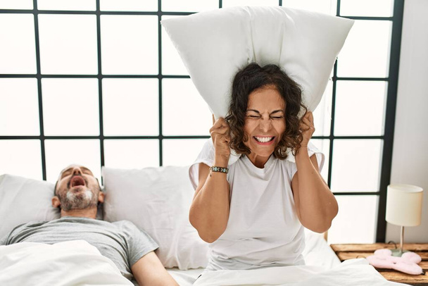 Middle age hispanic man snoring and woman angry covering ears with pillow at bedroom. - Foto, Imagen