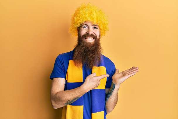 Vöröshajú férfi hosszú szakállú futball huligán éljenzés játék visel vicces paróka meghökkent, és mosolygott a kamerába, miközben bemutatta a kezét, és ujjal mutogatott.  - Fotó, kép