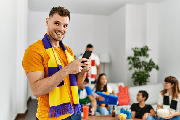 Ομάδα νεαρών φίλων που παρακολουθούν και υποστηρίζουν ποδοσφαιρικό αγώνα. Ο άνθρωπος χαμογελά χαρούμενος χρησιμοποιώντας smartphone στο σπίτι. - Φωτογραφία, εικόνα