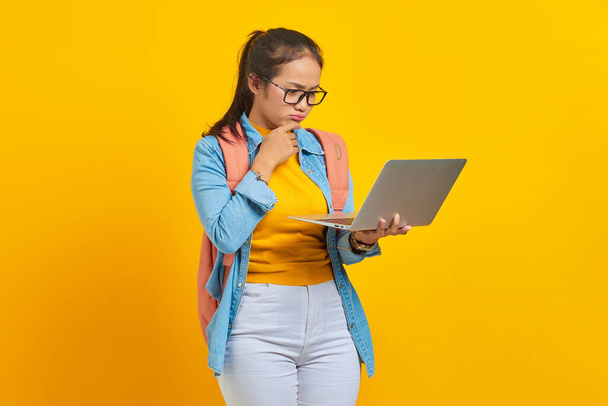 Portret poważnej młodej Azjatki uczennicy w luźnych ubraniach z plecakiem z laptopem i myślącej o pytaniu odizolowanym na żółtym tle. Edukacja w koncepcji uczelni wyższych - Zdjęcie, obraz