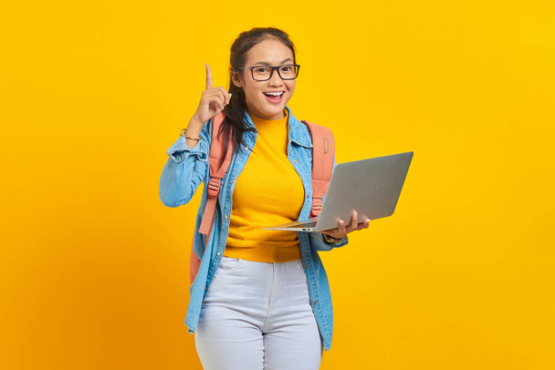 Portret wesołej młodej Azjatki uczennicy w ubraniach casual z plecakiem za pomocą laptopa i o kreatywnym pomyśle odizolowanym na żółtym tle. Edukacja w koncepcji uczelni wyższych - Zdjęcie, obraz