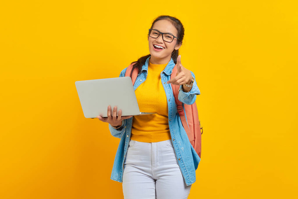 Portrait of cheerful young Asian woman student in casual clothes with backpack using laptop and pointing at camera with finger isolated on yellow background. Education in college university concept - Foto, imagen