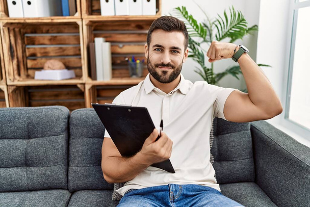 Handsome hispanic man holding clipboard working at psychology clinic strong person showing arm muscle, confident and proud of power  - Photo, Image