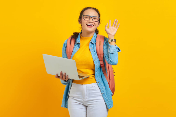 Retrato de una joven estudiante asiática sonriente vestida con ropa casual con mochila usando laptop y haciendo gestos con la mano aislada sobre fondo amarillo. Educación en concepto universitario universitario - Foto, Imagen
