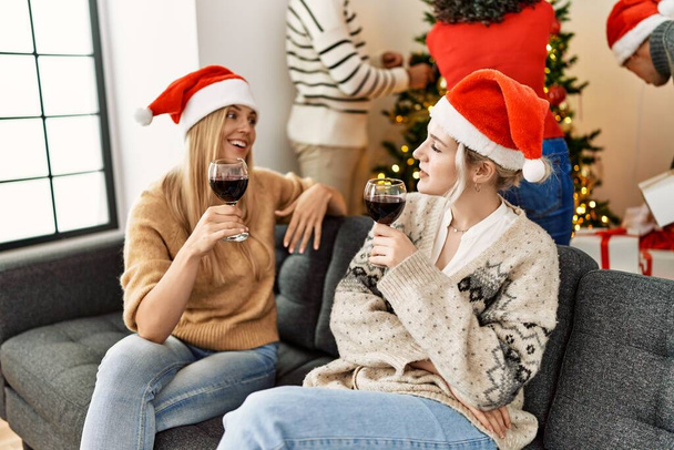 Group of young people celebrating christmas. Two woman speaking and toasting with glass of wine at home. - Photo, Image
