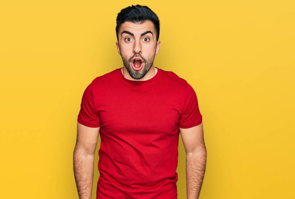 Hombre hispano con barba vistiendo camiseta roja casual asustado y sorprendido con la expresión sorpresa, el miedo y la cara emocionada.  - Foto, imagen
