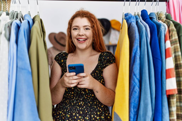 Молода руда жінка шукає одяг на стійці для одягу, використовуючи смартфон, який виглядає позитивно і щасливо стоячи і посміхаючись з впевненою посмішкою, показуючи зуби
  - Фото, зображення