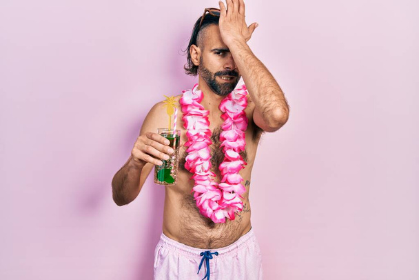 Jonge Spaanse man in badpak en Hawaïaanse lei drinken tropische cocktail verrast met hand op hoofd voor fout, herinner fout. vergeten, slecht geheugen concept.  - Foto, afbeelding
