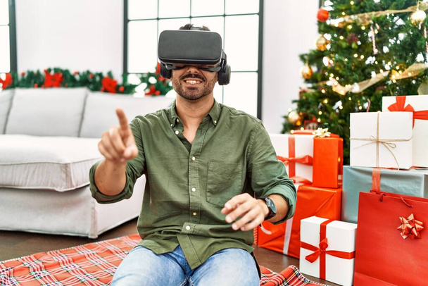 Genç İspanyol adam VR gözlük takıyor. Evdeki Noel ağacının yanında oturuyor. - Fotoğraf, Görsel