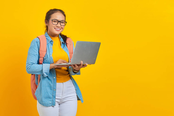 Retrato de jovem estudante asiática alegre em roupas casuais com mochila usando laptop e olhando para a câmera isolada no fundo amarelo. Educação no conceito de faculdade universitária - Foto, Imagem