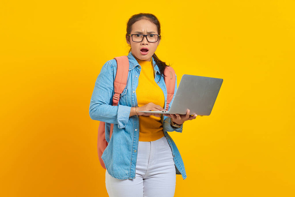 Портрет здивованої дівчини азіатського походження в повсякденному одязі з рюкзаком за допомогою ноутбука і погляд на камеру, ізольовану на жовтому тлі. Навчання в університетському коледжі - Фото, зображення