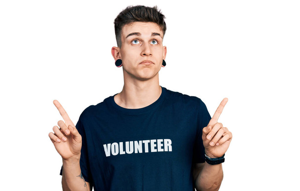 Молодий білий хлопчик з розширенням вух носить волонтерську сорочку, що вказує на сумний і засмучений вигляд, вказуючи напрямок пальцями, нещасний і пригнічений
.  - Фото, зображення