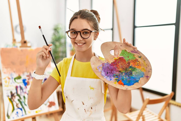 Rozkošná dívka usmívající se sebevědomě drží štětec a paletu v ateliéru umění - Fotografie, Obrázek