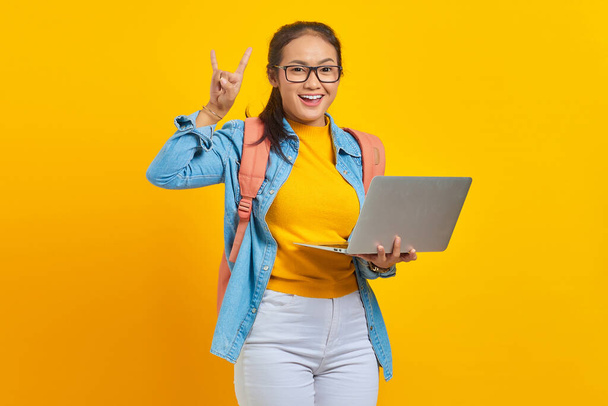 Retrato de jovem estudante asiática alegre em roupas casuais com mochila usando laptop e fazendo símbolo de rocha com as mãos para cima isolado no fundo amarelo. Educação no conceito de faculdade universitária - Foto, Imagem