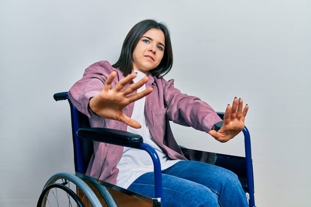 Young brunette woman sitting on wheelchair doing stop gesture with hands palms, angry and frustration expression  - Фото, зображення