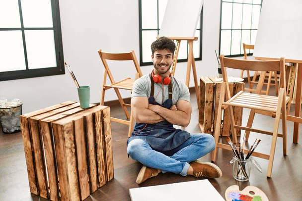 Young hispanic man sitting at art studio happy face smiling with crossed arms looking at the camera. positive person.  - Foto, imagen