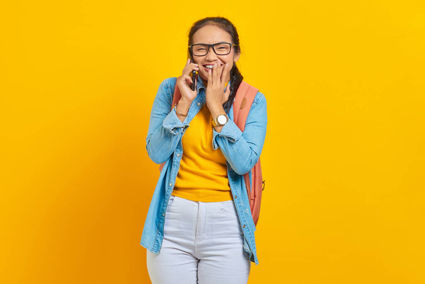 Portrait de jeune asiatique joyeuse étudiante en vêtements décontractés avec sac à dos tenant smartphone et couvrant la bouche avec des paumes isolées sur fond jaune. Éducation dans le concept d'université collégiale - Photo, image