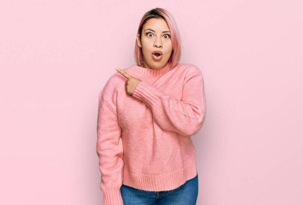 Hispánka s růžovými vlasy na sobě ležérní zimní svetr překvapeně ukazuje prstem na stranu, otevřená ústa užaslý výraz.  - Fotografie, Obrázek