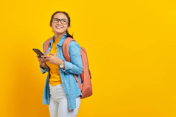 Ritratto di giovane studentessa asiatica sorridente in abiti casual con zaino utilizzando il telefono cellulare e guardando da parte isolata su sfondo giallo. Istruzione universitaria concetto di università - Foto, immagini