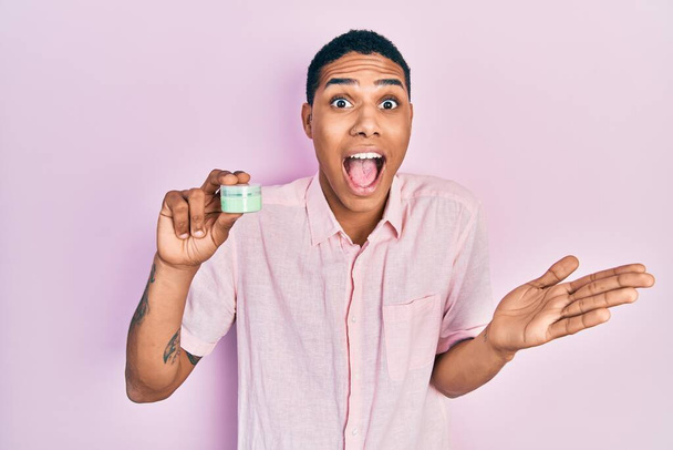 Young african american guy holding face moisturizer cream celebrating achievement with happy smile and winner expression with raised hand  - 写真・画像