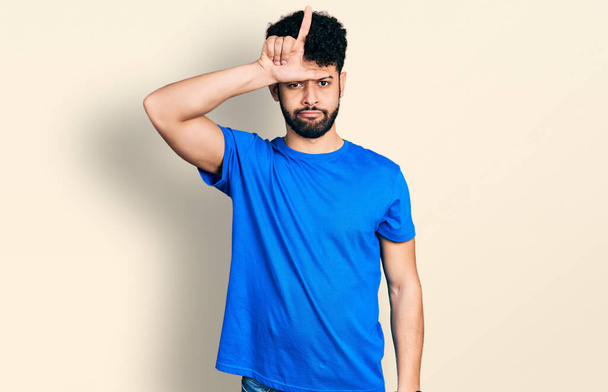 Young arab man with beard wearing casual blue t shirt making fun of people with fingers on forehead doing loser gesture mocking and insulting.  - Photo, Image