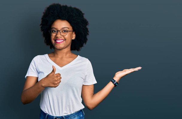 Fiatal afro-amerikai nő, alkalmi fehér pólóban, tenyérrel a kezében, feltartott hüvelykujjal, vidám mosollyal és vidám mozdulattal.  - Fotó, kép