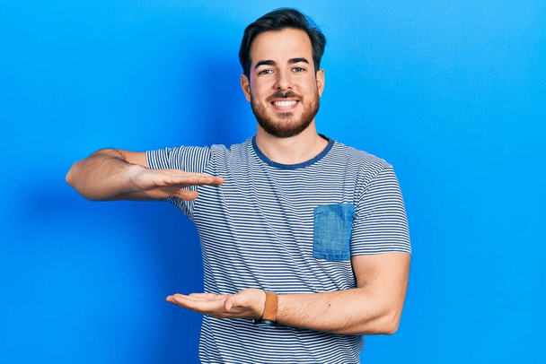 Handsome caucasian man with beard wearing casual striped t shirt gesturing with hands showing big and large size sign, measure symbol. smiling looking at the camera. measuring concept.  - 写真・画像