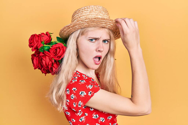 Beautiful caucasian woman with blond hair holding bouquet of red roses in shock face, looking skeptical and sarcastic, surprised with open mouth  - Zdjęcie, obraz