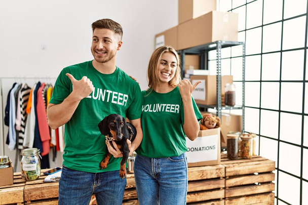 Fiatal pár aranyos kutya visel önkéntes póló adományok állni mutatva hüvelykujját felfelé mosolyogva boldog nyitott szájjal  - Fotó, kép