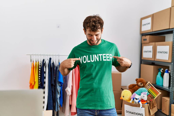 Young hispanic man smiling happy pointing with fingers volunteer uniform at charity center. - Photo, Image