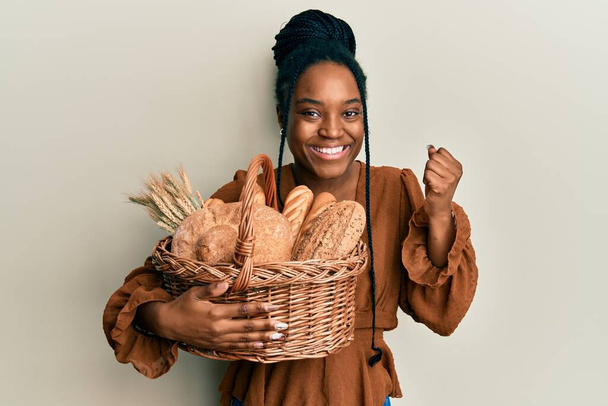 Mujer afroamericana con el pelo trenzado sosteniendo canasta de mimbre con el pan gritando orgulloso, celebrando la victoria y el éxito muy emocionado con el brazo levantado  - Foto, Imagen