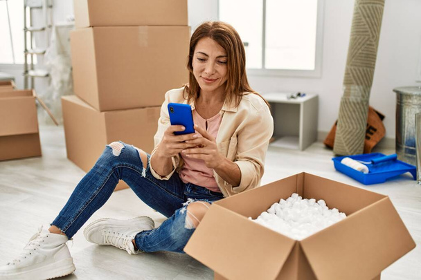 Biała kobieta w średnim wieku za pomocą smartfona siedzi na podłodze w nowym domu. - Zdjęcie, obraz