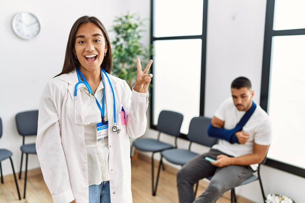 Fiatal ázsiai orvos nő a váróteremben egy törött karú férfival, aki a kettes számú ujjal mutogat, miközben magabiztosan és boldogan mosolyog..  - Fotó, kép