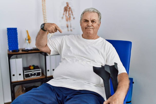 Uomo anziano caucasico alla clinica di fisioterapia che tiene le stampelle guardando fiducioso con il sorriso sul viso, puntandosi con le dita orgoglioso e felice.  - Foto, immagini