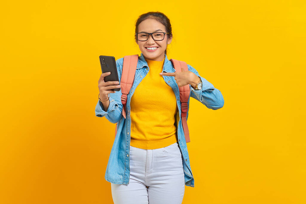 Portrét veselého mladého asijského studenta v neformálním oblečení s batohem ukazujícím na mobilní telefon s prsty izolovanými na žlutém pozadí. Vzdělání v univerzitní koncepci - Fotografie, Obrázek