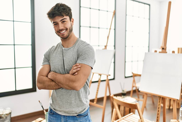 Молодой латиноамериканец улыбается счастливо стоя со скрещенными руками в художественной студии. - Фото, изображение