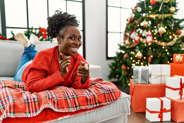 Fiatal afro-amerikai nő fekszik a kanapén karácsonyfán, ujjal mutogatva a kamerára boldog és vicces arccal. jó energia és rezgések.  - Fotó, kép
