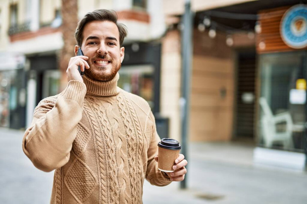 Νεαρός καυκάσιος άνδρας μιλάει στο smartphone πίνοντας καφέ στην πόλη. - Φωτογραφία, εικόνα