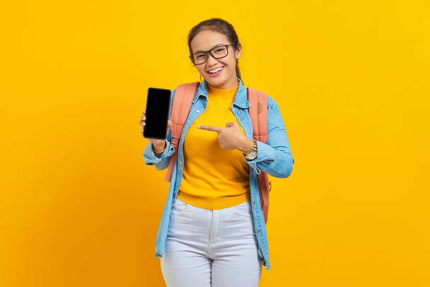 Πορτρέτο του χαρούμενα νεαρή Ασιάτισσα φοιτήτρια σε casual ρούχα με σακίδιο που δείχνει κενό οθόνη κινητό τηλέφωνο απομονώνονται σε κίτρινο φόντο. Εκπαίδευση στο πανεπιστήμιο έννοια - Φωτογραφία, εικόνα
