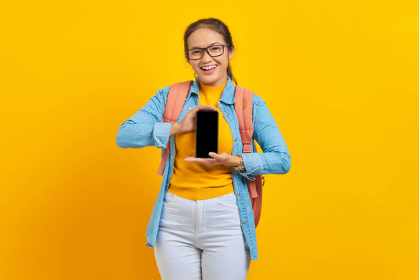 Portrét veselého mladého asijského studenta v neformálním oblečení s batohem ukazujícím prázdný displej mobilního telefonu izolovaného na žlutém pozadí. Vzdělání v univerzitní koncepci - Fotografie, Obrázek