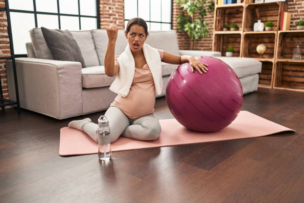 Молодая беременная женщина сидит на коврике для йоги с мячом для пилатеса раздраженный и разочарованный кричать от гнева, кричать сумасшедшие от гнева и рука поднята  - Фото, изображение