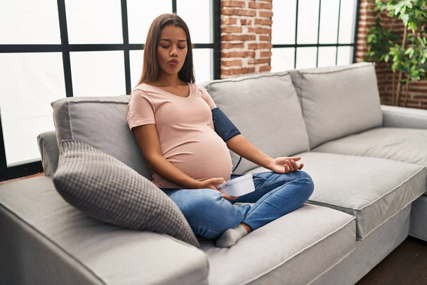 Fiatal terhes nő használ vérnyomás monitor ül a kanapén, hogy hal arcát a száj és hunyorgó szemek, őrült és komikus.  - Fotó, kép
